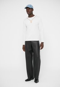 Biela košeľa s dlhým rukávom, zlatý reťazový náhrdelník, čierne kožené nohavice s širokými nohavicami a svetlomodrá čiapka s nápisom. Čierne topánky dopĺňajú outfit.