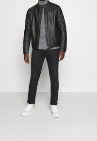 Man som bär en svart läderjacka, grå polotröja, svarta uppvikta byxor och vita sneakers som står mot en enkel bakgrund.