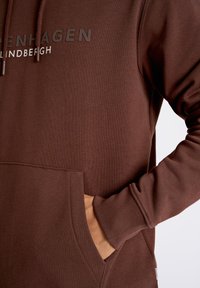 Brun huvtröja i mjukt tyg med känguruficka, snörning i huvan och tryckt text "KÖPENHAMN" i en kontrasterande färg.