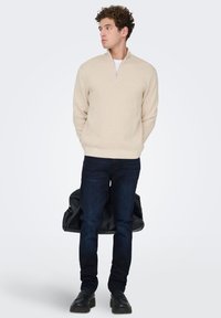 Beige zip-up sweater with long sleeves, paired with dark blue jeans and black shoes, holding a black duffel bag.