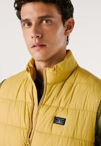 Gilet imbottito giallo con alto colletto, caratterizzato da un design a righe orizzontali e un piccolo etichetta del marchio sul petto. Chiusura frontale con zip.