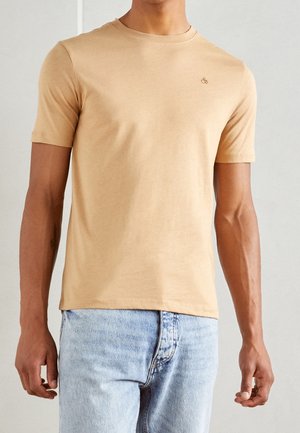 Man wearing a plain beige short-sleeve crew neck t-shirt with light blue jeans against a neutral background.