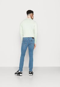 Homem com cabelo encaracolado curto, vestindo um suéter claro, jeans azuis e sapatilhas Nike Air pretas, posando contra um fundo branco simples.