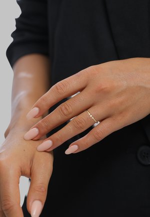 Rechte Hand mit gepflegten, blassrosa lackierten Nägeln und einem goldenen Ring mit drei kleinen Steinen am Ringfinger, der auf dem linken Handgelenk ruht, vor schwarzer Kleidung.