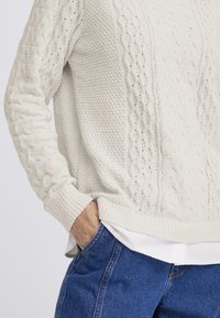 Maglione beige chiaro lavorato a maglia con motivi a rilievo e polsini a coste, abbinato a jeans blu. Il materiale sembra morbido e traspirante.