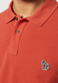 Gros plan d'une personne portant un polo orange rouille avec un patch zèbre rayé coloré sur la poitrine gauche et deux boutons marron.