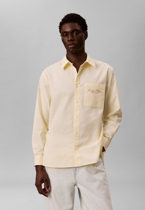 Mann trägt hellgelbes Hemd mit Knöpfen und einer vorderen Tasche mit dem "Calvin Klein"-Logo, kombiniert mit weißen Hosen vor einem einfarbigen grauen Hintergrund.