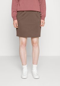 Hnědá sukňa z džerseje s bočními kapsami, spárovaná s růžovou mikinou a bílými teniskami. Design je přiléhavý a ke kolenům s jednoduchým lemem.