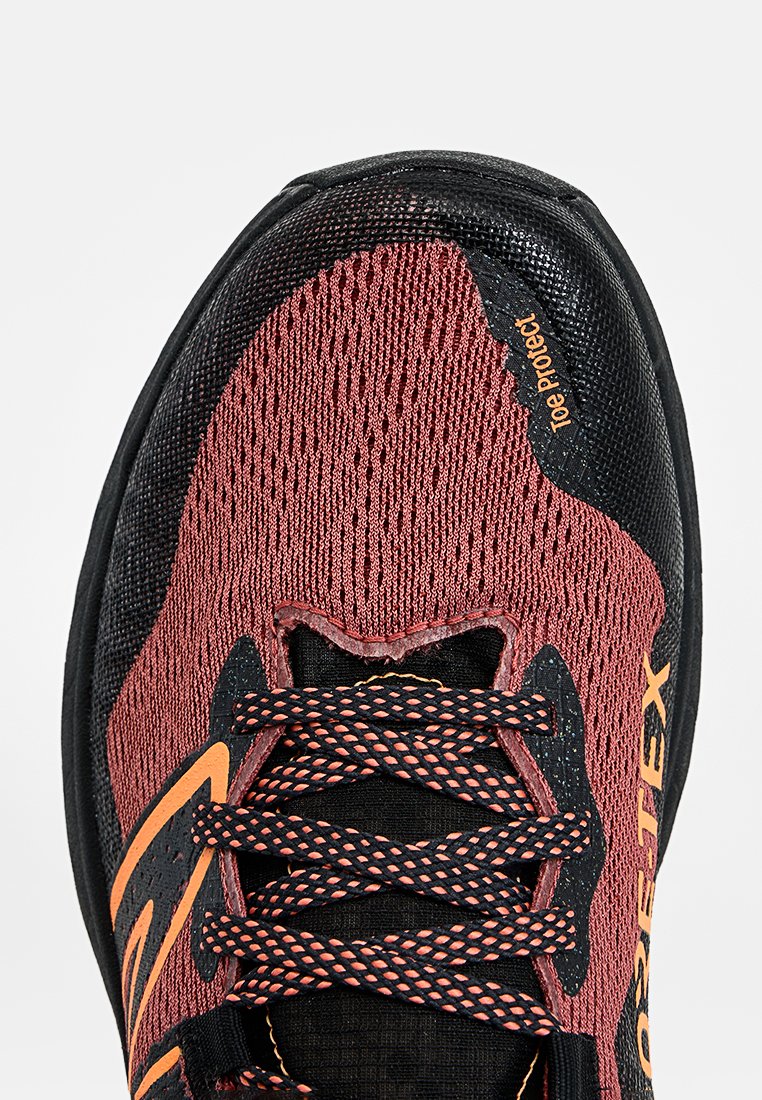 Primer plano de un zapato deportivo rojo y negro con acentos naranjas, cordones con patrón y etiqueta de "Protección de la puntera" en la zona delantera.