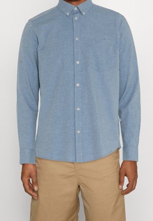 Chemise bleu clair à boutons avec manches longues, une poche sur la poitrine gauche, et un col. Portée avec un pantalon beige.