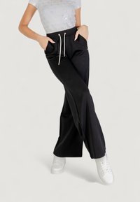 Pantalones negros de pierna ancha con un patrón texturizado, que cuentan con bolsillos laterales y una cintura con cordón. Combinados con zapatillas blancas.
