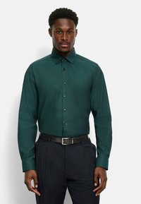 Camicetta button-up verde scuro con un motivo testurizzato, maniche lunghe, colletto a punta e bottoni neri, abbinata a pantaloni scuri e una cintura nera.