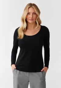 Schwarzes Langarmshirt mit Rundhalsausschnitt, aus weichem Stoff, kombiniert mit hellgrauen, maßgeschneiderten Hosen, die Hände in den Taschen.