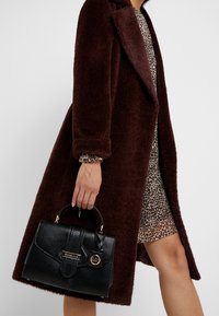 Svart texturerad handväska med en strukturerad design, guldfärgade detaljer och ett kort handtag. Bärs med en brun kappa och en leopardmönstrad klänning.