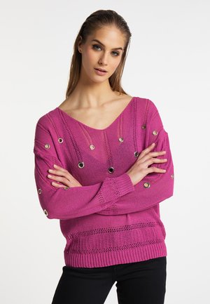 Jonge vrouw draagt een roze gebreide trui met metalen oogjes, staat met gekruiste armen tegen een effen witte achtergrond.