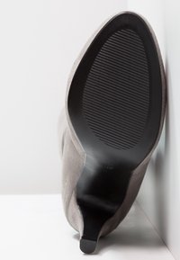 Szare zamszowe botki na wysokim obcasie, leżące na boku, ukazujące czarną teksturowaną podeszwę oraz smukły stężony obcas na białym tle.