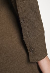 Tecido castanho, texturizado, com uma aparência rugosa. Apresenta uma manga com botão e dois botões escuros, destacando um detalhe de design bem cortado.