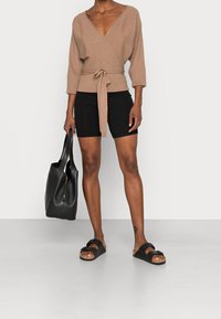 Bruin wikkelshirt met driekwartsmouwen en strik in de taille, gecombineerd met zwarte korte broek. Geaccentueerd met een zwarte tote tas en sandalen.