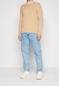 Beige neulepaita, jossa on resorireunus, yhdistetty vaaleansinisissä farkuissa ja valkoisissa tennareissa, esittelee rentoa istuvuutta ja minimaalisia merkintöjä.