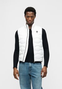 Άνδρας που φοράει ένα λευκό γιλέκο με καπιτονέ ύφασμα πάνω από μαύρο μπλουζάκι με μακριά μανίκια και μπλε τζιν, στέκεται μπροστά σε απλό φωτεινό φόντο.