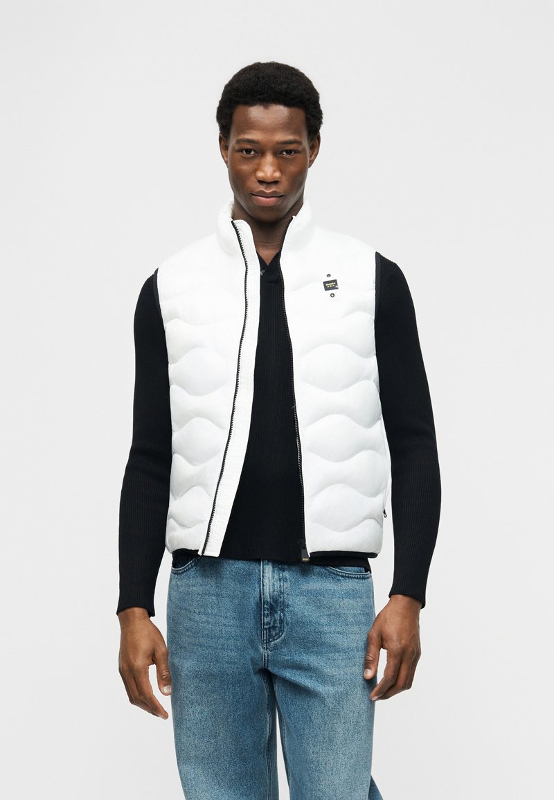 Άνδρας που φοράει ένα λευκό γιλέκο με καπιτονέ ύφασμα πάνω από μαύρο μπλουζάκι με μακριά μανίκια και μπλε τζιν, στέκεται μπροστά σε απλό φωτεινό φόντο.