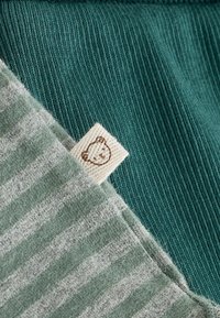 Baumwollmischgewebe mit einem türkisfarbenen gerippten Abschnitt und einem grauen gestreiften Patch. Ein kleines Stofflabel mit einer Bärenillustration ist sichtbar.