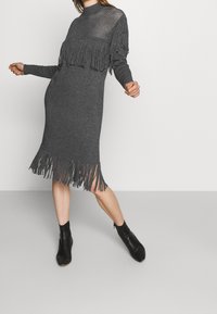 Femme portant une robe en tricot gris foncé avec des détails en frange sur la poitrine et l'ourlet, ainsi qu'un panneau en maille, associée à des bottines noires, sur fond uni.
