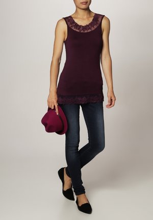 Femme portant un haut sans manches violet foncé avec bordure en dentelle, un jean skinny foncé, des ballerines noires, tenant un chapeau violet assorti.