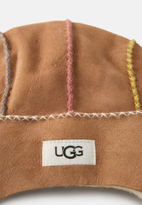 Ruskea samettihattu, jossa on värikkäitä ompeleita vaaleanpunaisella, sinisellä ja keltaisella. Varustettu valkoisella etiketillä, jossa lukee "UGG" mustalla langalla. Pehmeä rakenne.