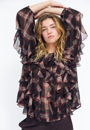 Mujer joven con pelo rubio ondulado que lleva una blusa de cuadros transparente con volantes, levantando los brazos por encima de la cabeza contra un fondo blanco liso.