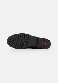 Semelle de chaussure en caoutchouc noir avec un motif de texture pour une adhérence, arborant un logo "Rieker" rouge et divers motifs de semelle pour une traction optimale.