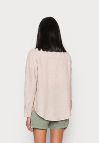 Lys beige linned skjorte med lange ærmer, der har en lige kant og sideslidser, parret med olivengrønne shorts. Simpelt design, glat tekstur.