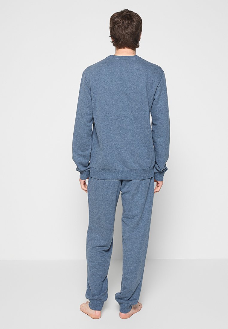 Personne debout pieds nus, de dos, portant un ensemble coordonné de haut à manches longues et de pantalons bleus sur un fond uni.