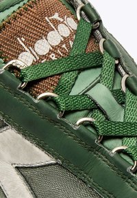 Sneaker verde con un tessuto marrone texture, occhielli metallici e lacci verdi a contrasto. Accenti in pelle vicino alla suola arricchiscono il design.