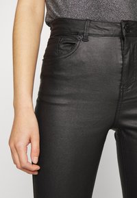 Närbild av en person som bär blanka svarta högmidjade byxor och en glittrig grå topp, med vänster arm och hand avslappnat vid sin sida.