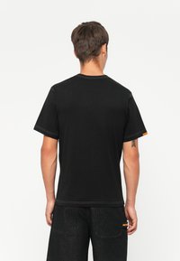 Μαύρη βαμβακερή μπλούζα με κοντά μανίκια, στρογγυλή λαιμόκοψη, αντίθετη ραφή και μια μικρή πορτοκαλί ετικέτα στη πλευρά.