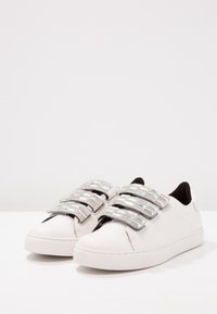 Baskets en cuir blanc à la texture lisse, avec quatre sangles métalliques scintillantes sur le dessus et une semelle en caoutchouc.