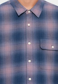 Button-up shirt in a blue and pink plaid pattern, featuring a chest pocket, a classic collar, and white buttons on a textured fabric.