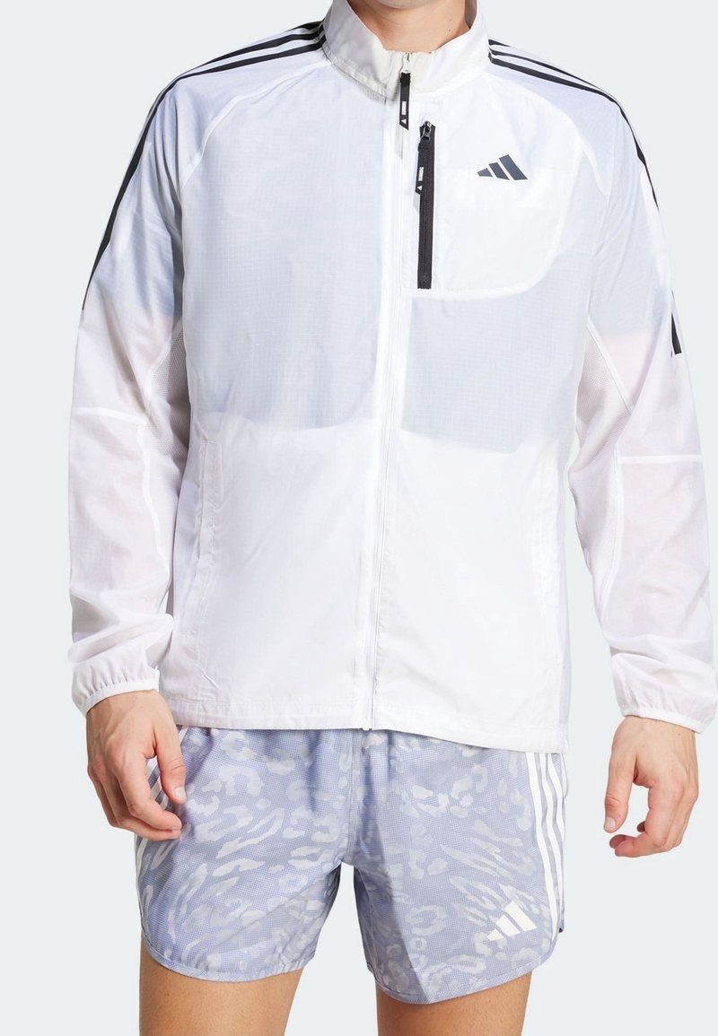 Homme portant une veste de sport blanche à fermeture éclair avec un logo noir et un short de sport gris clair à motif avec des bandes blanches.