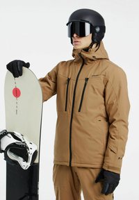 Chaqueta de esquí impermeable marrón con dos bolsillos con cremallera, acompañada de una tabla de snowboard y fijaciones blancas. Casco negro brillante y gafas de esquí.