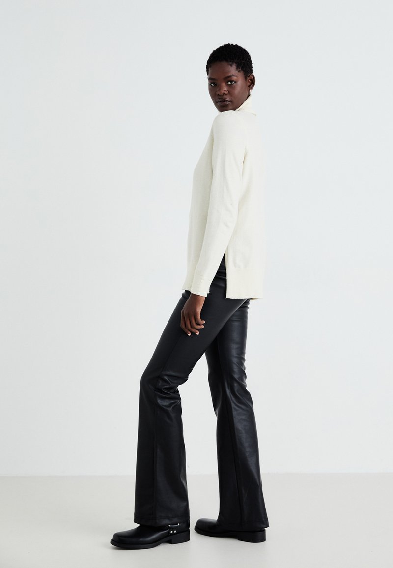 Maglione crema con collo alto e spacchi laterali, abbinato a pantaloni finti in pelle nera a zampa d'elefante e stivali neri chunky. Design semplice e moderno.