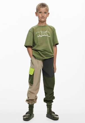 Zöld póló fehér grafikával és felirattal, bézs és zöld többpaneles cargo nadrág oldalzsebbel és kötött szegélyekkel, fekete szandál.