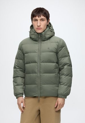 Jeune homme portant une veste matelassée à capuche verte et un pantalon beige, debout face à l’avant avec une expression neutre.