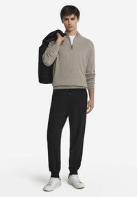 Jasnoszara bluza z zamkiem na suwak, czarne dresowe spodnie z kieszeniami oraz białe sneakersy. Model trzyma czarną pikowaną kurtkę na jednym ramieniu.