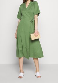 Abito verde avvolgente con maniche corte, gonna di lunghezza media, texture morbida e una cintura verde coordinata. Abbinato a una borsa clutch beige e sandali bianchi.