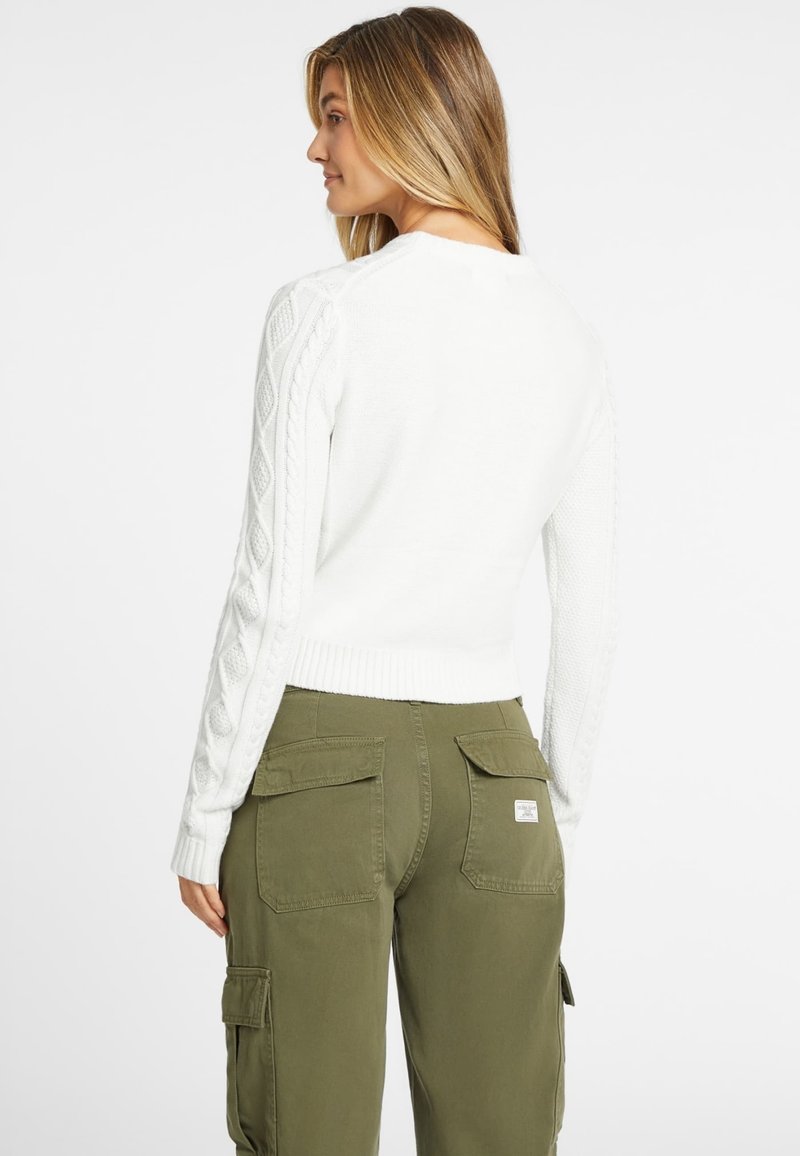 Weißer Strickpullover mit Rippenbund und -ärmeln, kombiniert mit olivgrünen Cargo-Hosen mit zwei Gesäßtaschen.