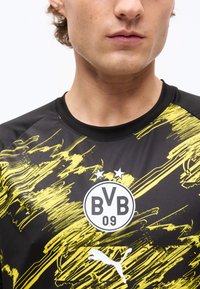Κοντινή εικόνα ενός ατόμου που φοράει μαύρη και κίτρινη φανέλα ποδοσφαίρου της Borussia Dortmund με το λογότυπο BVB 09 και το λευκό λογότυπο της Puma.