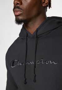 Hoodie noir en mélange de coton, arborant un logo "Champion" brodé en noir. Il possède une capuche avec cordon de serrage et une finition texturée.