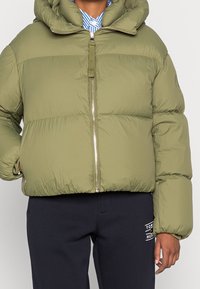 Osoba ubrana w oliwkową kurtkę puchową z zamkiem z przodu oraz granatowe spodnie z małym białym logo na udzie.