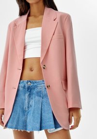 Žena nosí oversized růžový blejzr přes bílý bandeau top a plisovanou modrou džínovou minisukni s třásněmi.
